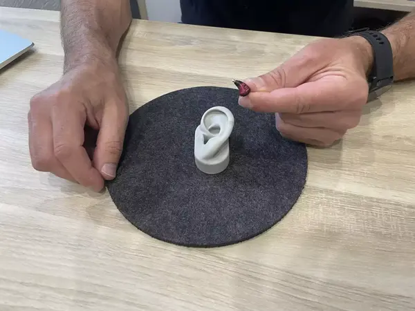 Ein Mann zeigt ein kleines Hörgerät neben einem Ohrmodell auf einem Holztisch.